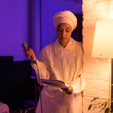 Eine Person in heller Kleidung und Kopftuch lehnt an einer Wand neben einer Lampe. In der Hand hält sie ein Mikrofon und ein Heft, aus dem sie vorzulesen scheint.