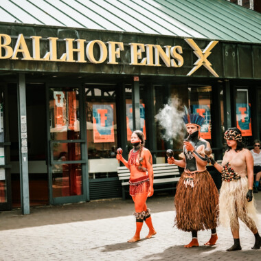 Three people in traditional attire and body paint during a procession in front of "Ballhof Eins.