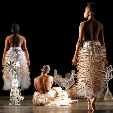 Ein Foto auf dem Stück Hatched Ensemble. Die Rückenansicht von drei Tänzer*innen. Sie sind Oberkörperfrei und tragen lange weiße Röcke aus Tüll, die mit hölzernen Wäscheklammern versehen sind. 