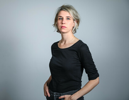 The photo shows Melanie Zimmermann in front of a grey background. She wears a black top with 3/4 sleeves, jeans and a discreet silver necklace. Her chin length, blonde hair ist styled with a deep side parting. She has her hands in her pants pockets and looks directly into the camera.