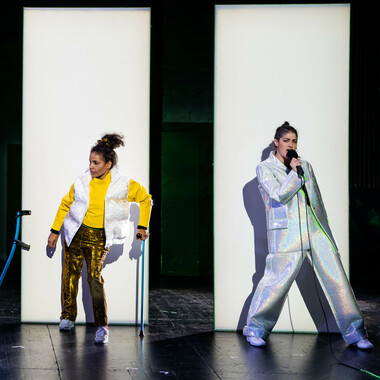Two people are standing next to each other, a little distance apart, each in front of a white high partition. The person on the left is wearing gold-coloured trousers, white sneakers, a yellow top and a white waistcoat. Her dark curly hair is tied in a braid. She leans on a blue crutch with her left hand and looks to her right. To her right, the second crutch leans against the partition wall.  On the right stands the second person, fully dressed in a loose silver reflective suit. She has dark hair, also tied in a braid, holds a microphone in front of her mouth and sings. Her hip is slightly bent, she leans back a little.