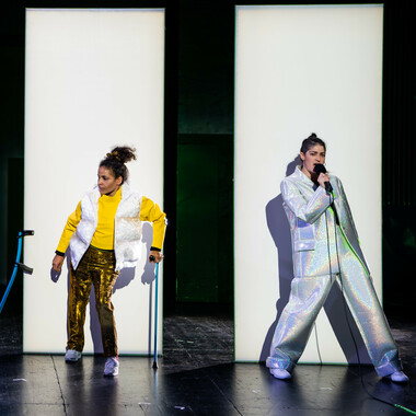 Two people are standing next to each other, a little distance apart, each in front of a white high partition. The person on the left is wearing gold-coloured trousers, white sneakers, a yellow top and a white waistcoat. Her dark curly hair is tied in a braid. She leans on a blue crutch with her left hand and looks to her right. To her right, the second crutch leans against the partition wall.
On the right stands the second person, fully dressed in a loose silver reflective suit. She has dark hair, also tied in a braid, holds a microphone in front of her mouth and sings. Her hip is slightly bent, she leans back a little.