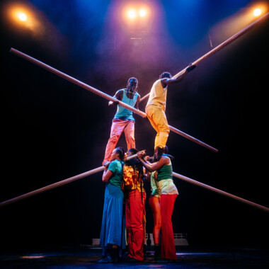 Eine Gruppe von Artist:innen in bunten Kostümen auf der Bühne. 4 bilden die Basis, 2 stehen auf deren Schultern. In ihren Händen halten sie lange Stangen. Die Szene ist von farbigem Bühnenlicht beleuchtet. 