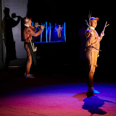 A stage immersed in pink and blue light. Ivor MacAskill stand on the right. Ivor wears a brown costume with a wooden look and a cap on his head. Ivor looks confused and holds up two wooden branches with both hands. Rosana Cade wears the same costume can be seen in the background. Rosana is standing in front of a TV that shows a live image of Ivor, in which he covers his left ear with one hand. With their right hand, Rosana holds a microphone with a plush rectangular headpiece towards their mouth. They are wearing a black apron around their hips that hold wooden sticks in different lengths.