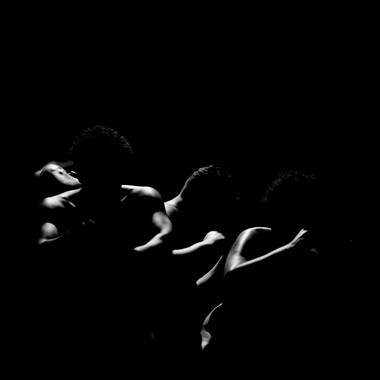 Image description by the editorial staff of Festival Theaterformen. Self-description of the depicted persons follows. / A black and white photo in front of a black background. In the center there are naked shoulders and arms of the performers, that are being spotlighted. 