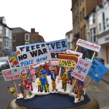 Kleine Figuren auf einem Sockel in der Innenstadt, die Protestschilder halten. Darauf steht unter anderem: "NO WAR PEACE NOW", "LOVE" oder "GIVE PEACE A CHAN`CE".