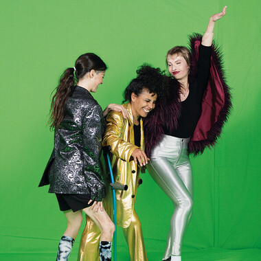 In the picture are three women: Alice Giuliani, Victoria Antonova and Laila White (from left to right), standing in front of a neon green background. Alice Giuliani is turning her back to the camera. Victoria Antonova is wearing a gold suit and leaning on a crutch, laughing. Alice Giuliani's hand is on her shoulder. On the far right is Laila White making a dance movement towards Victoria Antonova, wearing a cape with feathers and silver trousers.