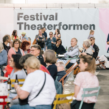 Eine lebendige Szene bei einem Festival im Freien mit vielen Menschen, die sich unterhalten, essen oder trinken. Im Hintergrund ist ein großes Banner mit der Aufschrift „Festival Theaterformen“ zu sehen. Die Veranstaltung findet zwischen einem roten Backsteingebäude und einem modernen weißen Gebäude statt. Es gibt bunte Infostände, Sitzgelegenheiten und eine entspannte Atmosphäre.