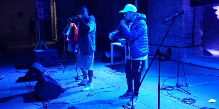 Das Musikduo Eskina Qom live auf einer Bühne, die in blaues Licht getaucht ist. Beide halten jeweils ein Mikrofon in der Hand und sind sportlich gekleidet. 