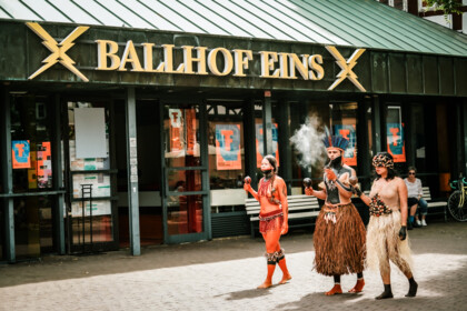 Lilly Baniwa, Ziel Karapotó und Olinda Tupinambá auf dem Ballhofplatz. Sie tragen auffällige Kostüme, im Hintergrund ist der Schriftzug Ballhof Eins.