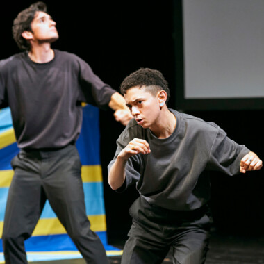 Two people in dark clothing on a stage in dynamic poses. The person in the foreground appears focused and seems to be performing a combative movement. In the background, a fabric in shades of blue and yellow is visible.