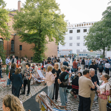 Eine große Gruppe von Menschen versammelt sich im Freien auf einem gepflasterten Platz zwischen Bäumen und Gebäuden. Die Stimmung ist lebendig und gesellig – viele Personen stehen in Gruppen, unterhalten sich oder trinken. Im Hintergrund sind ein rotes Backsteingebäude und ein modernes weißes Gebäude zu sehen. Einige bunte Klappstühle und Liegestühle stehen verteilt auf dem Platz, was dem Ort ein entspanntes Ambiente verleiht.