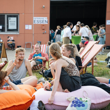 Mehrere Besuchende sitzen auf bunten Sitzsäcken und unterhalten sich auf einer Wiese vor einem Gebäude. Weitere Menschen stehen vor dem Eingang und kommen in kleinen Gruppen ins Gespräch.