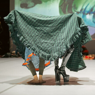 Stage situation. Two people under a large patterned cloth, only their legs and feet are visible. One person is barefoot, one is wearing high, laced leather boots.