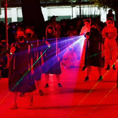 An open-air square immersed in red light. Four masked people in knee-length, dark capes stand one behind the other. With their right hands stretched forward, they operate green laser pointers. Behind them are two people, one in a yellow and the other in a red jumpsuit. Both are holding spotlights in their hands, but only the left one is being used. The event on the square is observed by a large number of people in the background of the image.