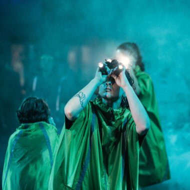Mehrere Personen in gelben Regenponchos stehen im dichten Nebel. Eine Person hält zwei Taschenlampen vor die Augen, als würde sie durch ein Fernglas blicken. Die Szene ist bläulich beleuchtet.