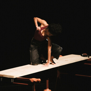 Bühnensituation: Eman Hussein kniet auf einem Holzbrett, das auf einem Gerüst liegt und scheint mit den Händen etwas zu hämmern.