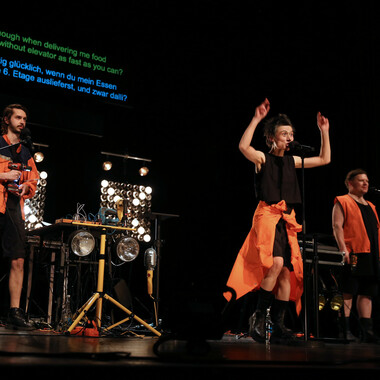 Image description by the editorial staff of Festival Theaterformen. Self-description of the depicted persons follows. / Three people in black and neon orange clothing are spread out on the stage, each holding a microphone. They are facing the audience. Green and blue text is projected in the background.