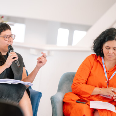 Two person sit together, one speaks into a microphone while the other looks at a notebook.