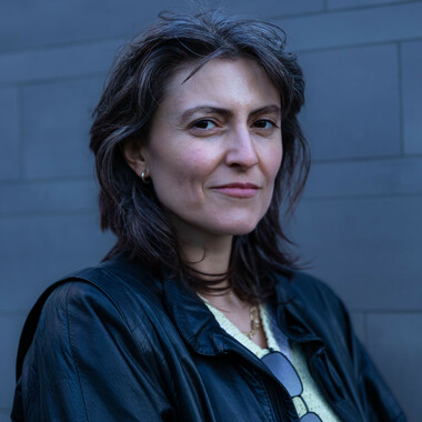 A portrait photo of Rita Mazza. Rita has black, shoulder-length hair with grey highlights. Rita is wearing a white T-shirt, a black leather jacket over it and has sunglasses tucked in front of the T-shirt. Rita is standing in front of a grey wall.