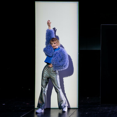 A person with a blond bob in front of a white high partition. She is wearing silver bell-bottom trousers and white sneakers, along with a blue top and a fluffy blue jacket. The person has her hips bent in and her right arm stretched up in the air. She holds her left hand to her stomach. She is looking into the audience.