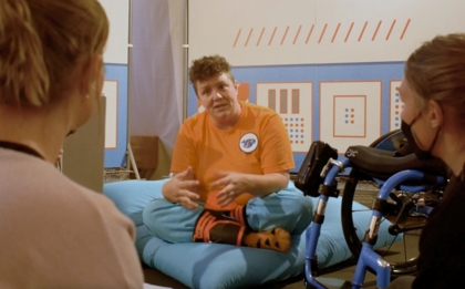 Jess Thom sitzt in einem blauen Sitzsack und spricht mit zwei Personen, die vor ihr sitzen. Rechts neben Jess Thom ist ein Rollstuhl zu sehen. Im Hintergrund sind Stellwände aufgebaut, die das "Raumschiff Biskuit" zeigen.