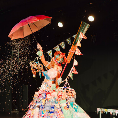 Person steht auf einer Bühne. Sie trägt ein buntes, aus recycelten Materialien gefertigten Kostüm mit großem Rock und vielen dekorative Objekte an denen viele Wimpelgirlanden befestigt wurden. In einer Hand hält sie einen roten Regenschirm. 