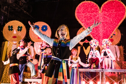 Stage setting: Many cheerful people with colourful traditional clothing made of LGBTQIA+ Pride flags, in the background dolls made of straw and a huge heart.