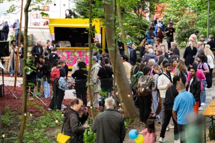Das Festivalzentrum am Gartenhaus Haeckel tagsüber. Es ist gefüllt mit Besucher*innen. Das Foto zeigt Festivalbesucher*innen. Sie stehen in Gruppen und reden miteinander. Einige halten Getränke in der Hand. Auf dem Platz eine gelbe Bude, die mit pinken Ko’eyene-Postern beklebt ist.