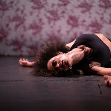 Ein Schwarzweiß-Foto aus dem Stück “When I saw the Sea” von Ali Chahrour: Eine Performer*in mit dunklen, schulterlangen, lockigen Haaren an einem Marmortisch, die in die Kamera blickt. Auf der Marmorplatte vor ihr ist die Reflektion der Person zu sehen.
