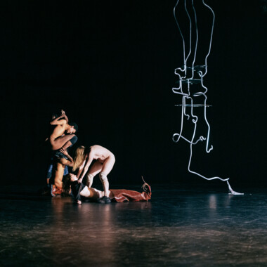 Mehrere nackte Performer bilden eine dichte, ineinander verschlungene Gruppe am Boden einer dunklen Bühne. Körper sind übereinander geschichtet und in einer skulpturalen Pose miteinander verbunden. Rechts im Bild hängt eine abstrakte, linienartige Skulptur von der Decke herab.