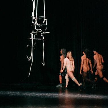 Vier nackte Performer bewegen sich gemeinsam über eine dunkle Bühne. Sie gehen von rechts nach links in einer Reihe, während über ihnen eine abstrakte, hängende Skulptur aus dünnen, hellen Stäben schwebt. 