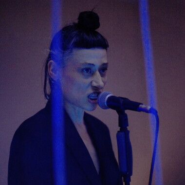 Eine Person mit ausdrucksstarkem Gesichtsausdruck spricht in ein Mikrofon. Blaues Licht und vertikale Lichtstreifen erzeugen eine kühle, intensive Bühnenatmosphäre.