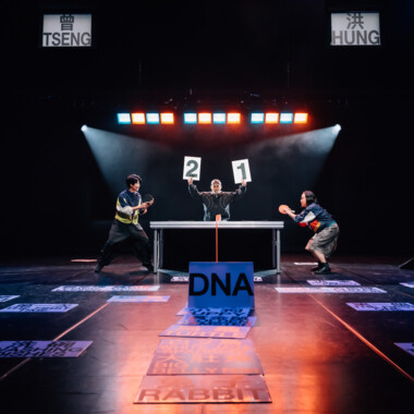 Drei Personen stehen auf einer Bühne um einen Tisch. Zwei weitere spielen Tischtennis, während die dritte Person Punktetafeln mit den Zahlen „2“ und „1“ hochhält. Im Vordergrund steht ein Schild mit der Aufschrift DNA auf dem Boden, umgeben von vielen weiteren beschrifteten Karten, die nicht zu lesen sind. 