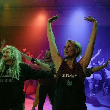 A club room immersed in colorful light, in which party guests can be seen dancing. They are  stretching their arms wide and looking to the left. The person in the foreground is wearing a black sleeveless top and has blond short hair.