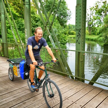 Jemand fährt mit einem Fahrrad über eine Brücke. Am Fahrrad ist ein Anhänger mit Solarpanels.