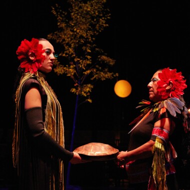 Zwei Personen stehen sich auf einer Bühne gegenüber. Sie halten ein Objekt zwischen sich, dass an einen Schildkrötenpanzer erinnert. Sie tragen jeweils eine große rote Blume im Haar. Warmes Licht beleuchtet die Szene, im Hintergrund ist ein Baum.