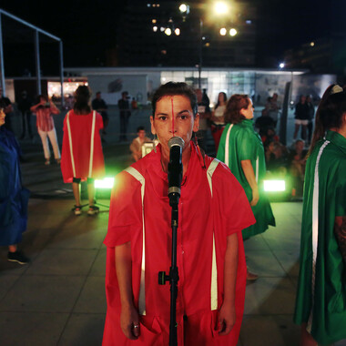 An open-air square illuminated with green spotlights at night. In the center of the image is a person in a red cape and feather earrings, standing upright in front of a microphone. The person appears to be speaking into it. The face is painted with a red line running from forehead to upper lip. The person's temples are painted with red dots. Behind the person, several people in red, green and blue capes can be seen. Their bodies are facing towards the right. The event on the square is observed by spectators in the background of the picture.