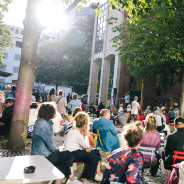 Menschen sitzen im Festivalzentrum und die Sonne scheint. Eine Lichterkette hängt zwischen zwei Bäumen.