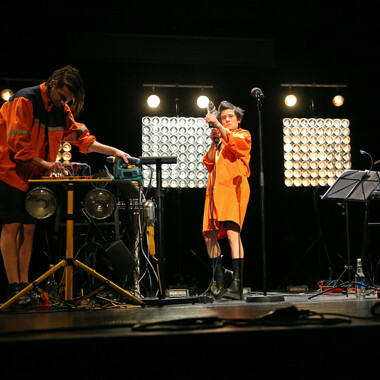 Image description by the editorial staff of Festival Theaterformen. Self-description of the depicted persons follows. / Two people in neon orange clothes are standing on the stage. Various tools and illuminants are distributed on the stage. The right person holds a drill with both hands and facing the audience. The left person is also handling different tools.