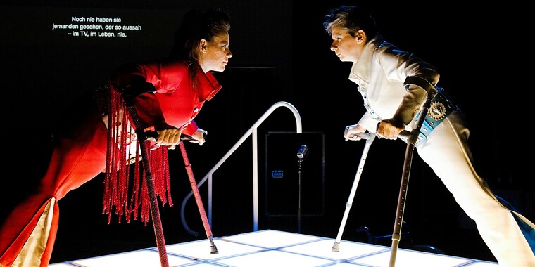 Two people on an illuminated platform facing each other. With their upper bodies bent far forward, they lean on their crutches and maintain intense eye contact. The person on the left wears a red jumpsuit with a wide belt, bell-bottoms and embellishments. She has dark, long hair combed back into a high ponytail. The person on the right is dressed in a white jumpsuit with a wide belt, flared pants and embellishments. The person has dark blonde, short hair.