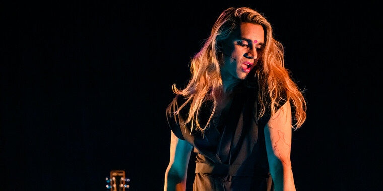 Self-description of the depicted person: A brown person wearing fuschia lipstick and a matching bindi, and a black jumpsuit with blond voluminous hair. Her head is tilted down to her left side. Attached to her cheek there is a small black microphone. In the background a small part of a guitar is visible.