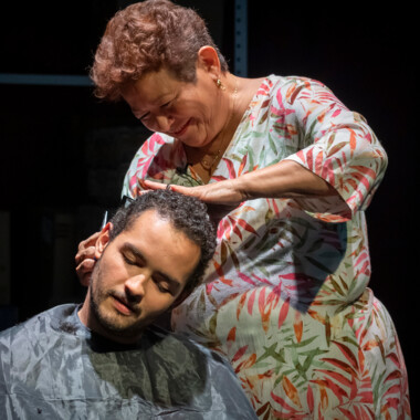 Eine ältere Frau schneidet einem jungen Mann die Haare. Sie trag ein bundgemustertes Kleid und lächelt. Er trägt einen Friseurumhang und hat die Augen geschlossen.